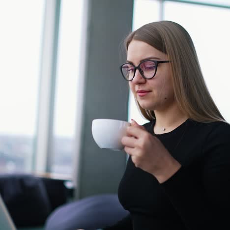 attractive woman drinking tea in office. portrait of a businesswoman in stylish glasses with a cup of coffee working on a laptop in the light office room