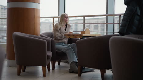 young lady sipping tea from her cup, dropping it on the table while looking outside through transparent glass in mall, with winter jacket hanging on chair