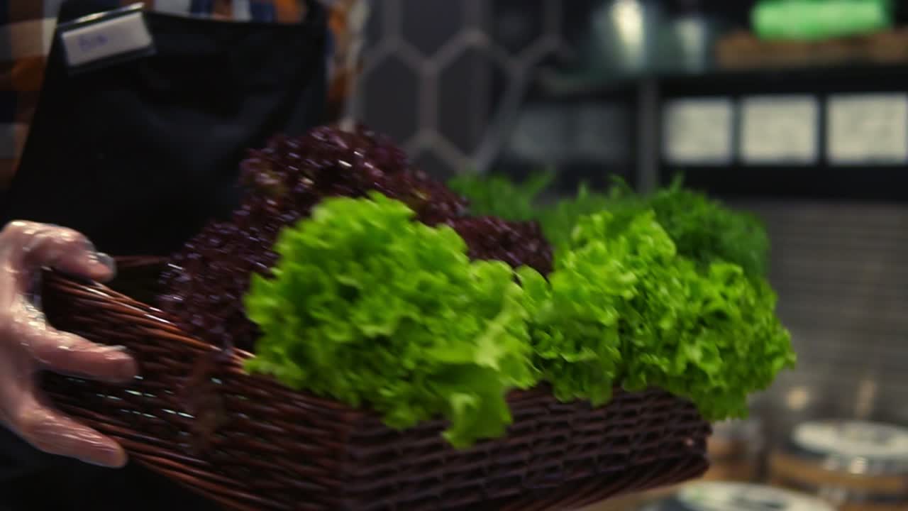 Seller man in apron in supermarket walking by vegetables aisle with box of fresh greens to arrange. Caucasian worker in local supermarket holding box of greens. Close up