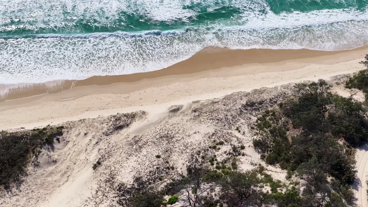 High angle drone shot pans from green scrubland across sand dunes to ocean waves