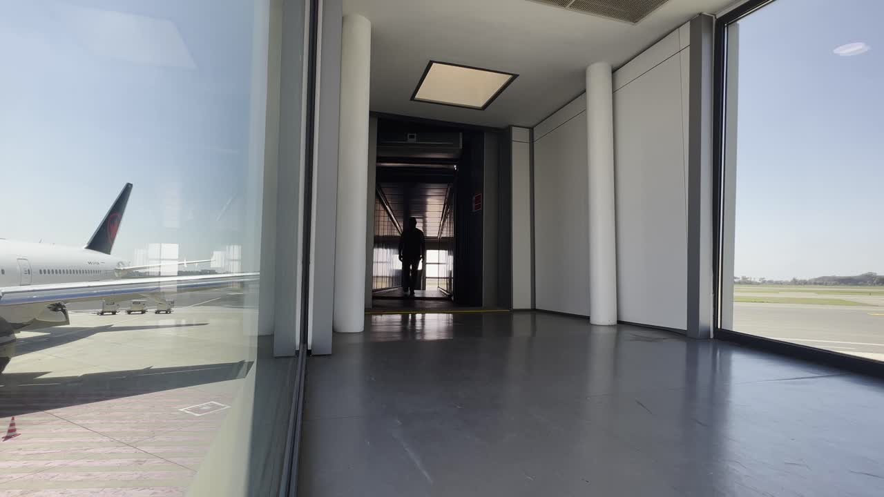 Airport worker in uniform walks towards the camera through a modern jet bridge, with an airplane visible on the tarmac through the window. Travel and aviation concept