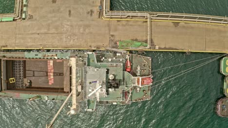 a large empty cargo ship with cranes docked at a concrete pier next to a group of tugboats