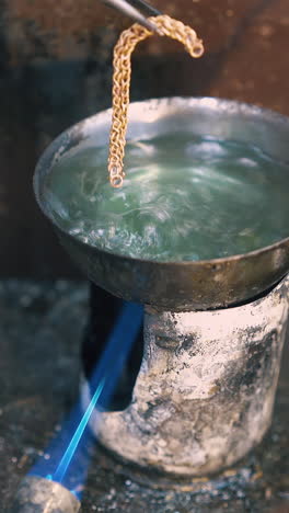 cu: skilled goldsmith takes gold chain off boiling green liquid on table near metal sheet in jeweler workshop closeup
