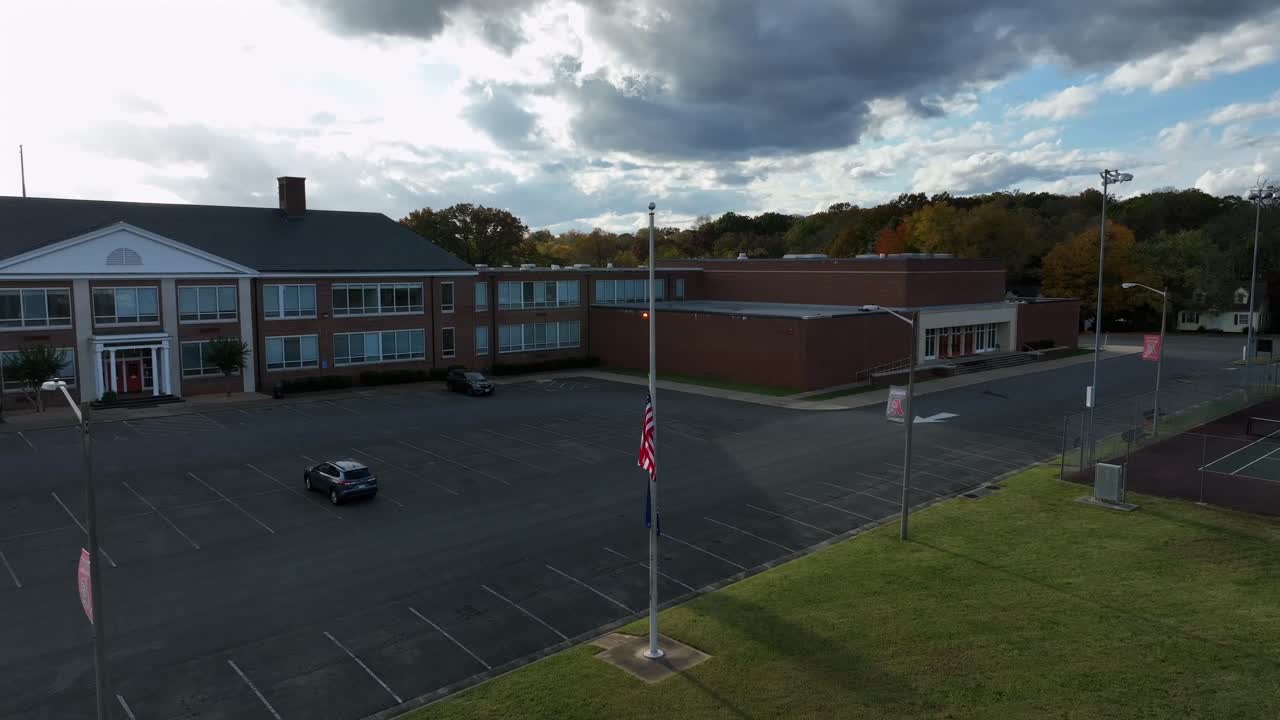 Aerial approaching shot of american flag on half mast on parking area of high school. Sunny day with red brick building during cloudy day in Virginia.