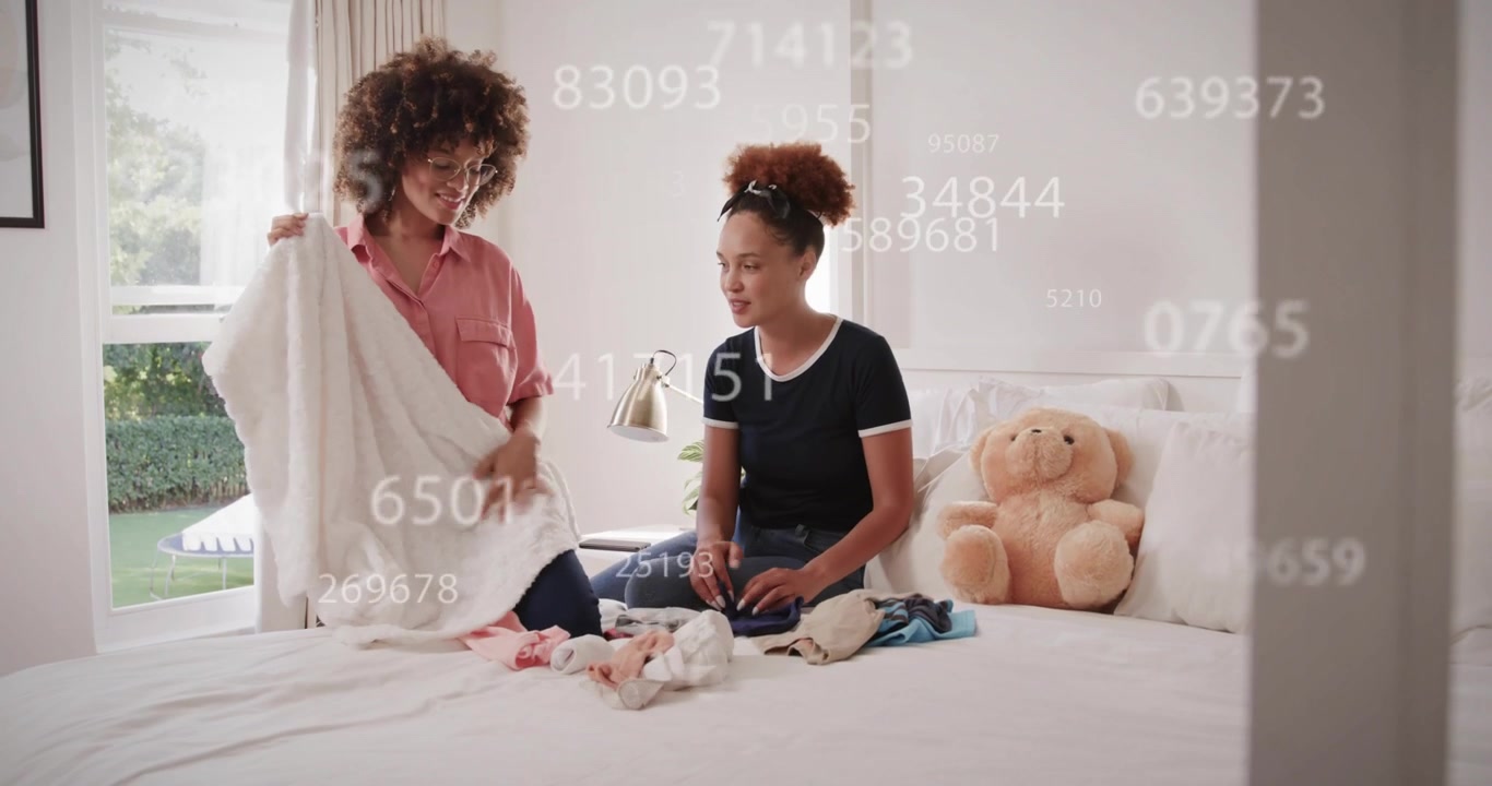 Standing woman carrying baby items and handing clothes to friend sorting on bed for nursery prep