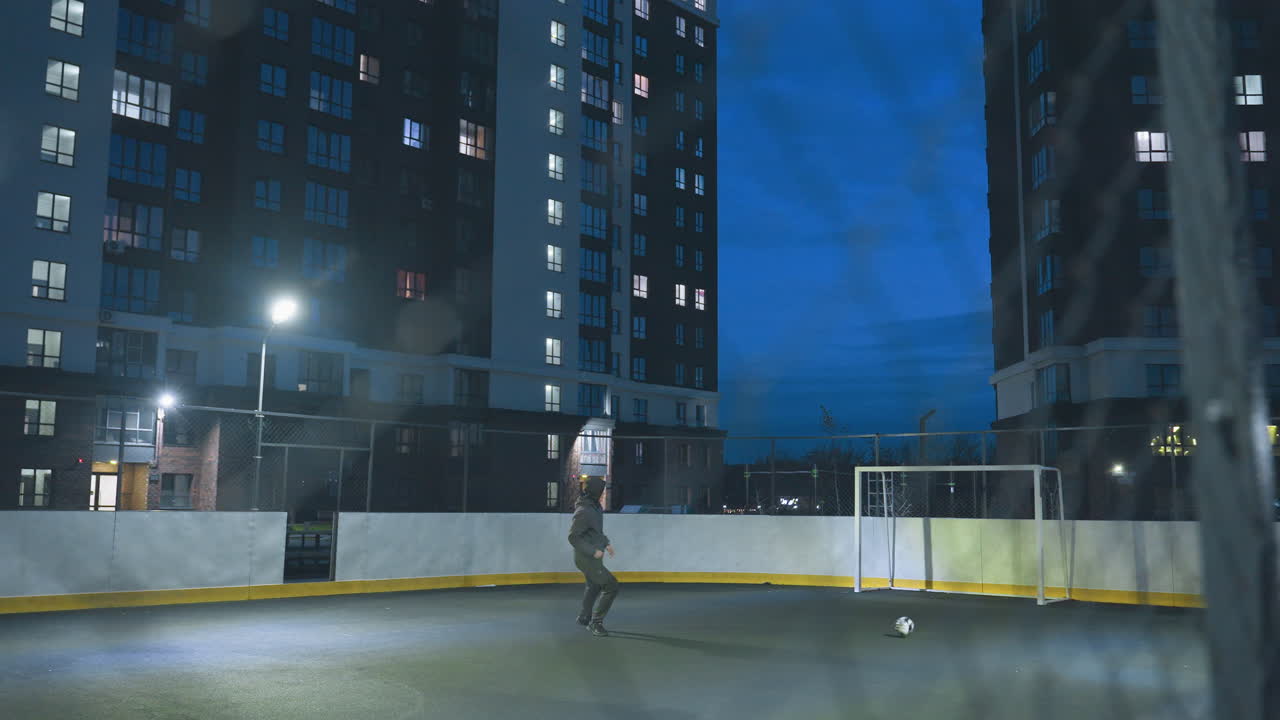 Soccer player kicks ball toward goalpost under bright urban lights, seen through entrance on outdoor field surrounded by tall residential buildings against a vibrant blue evening sky