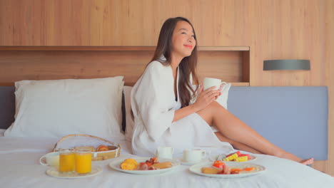 흰 가운을 입은 아시아 여성은 과일을 많이 곁들인 침대에서 아침 식사를 하면서 커피를 즐깁니다.