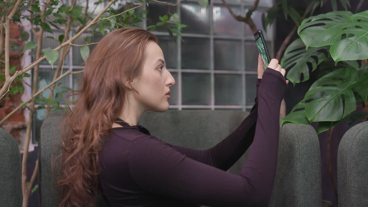 Pretty woman holding smartphone capturing close-up of green leaf while standing near cushioned chairs with brick wall and glass panel background in indoor plant-filled environment
