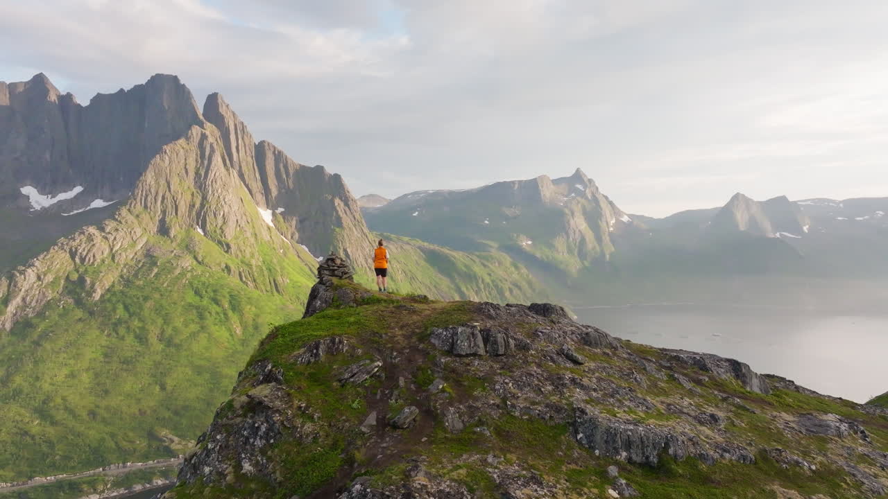 Hiker in arctic Senja overlooking jagged fjord mountain in sunset; drone reveal