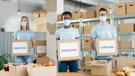 Grupo-Multiétnico-De-Voluntarios-Con-Máscara-Facial-Sosteniendo-Cajas-De-Donación-Y-Mirando-A-La-Cámara-En-Un-Almacén-De-Caridad