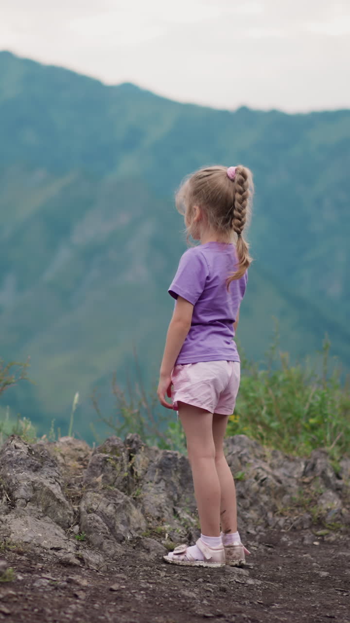 una niña pensativa con una camiseta púrpura y pantalones cortos mira las siluetas de las viejas cadenas montañosas en las tierras altas en un día sombrío vista trasera en cámara lenta