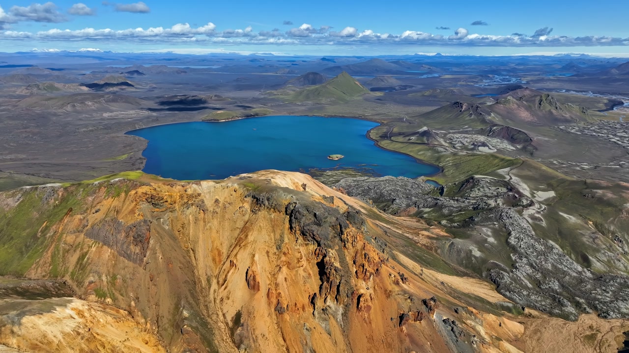 Sweeping drone panorama over a vivid blue volcanic lake and colorful rocky highlands under scattered clouds, showcasing remote wilderness and rugged terrain.