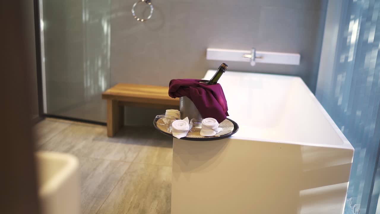 una toma lenta de una botella de champán romántica y vasos en una bandeja en el baño del hotel, con una gran bañera y una ventana en la escena.