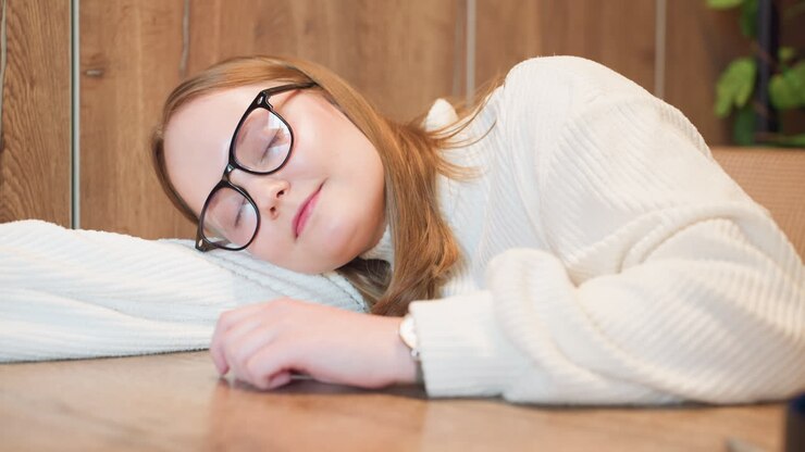 young girl in white sweater rests head gently on wooden table with eyes open and calm expression suggesting moment of pause, relaxation or quiet reflection in peaceful indoor setting