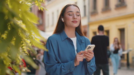 happy young woman in wireless earphones choosing listening music in smartphone dancing outdoors
