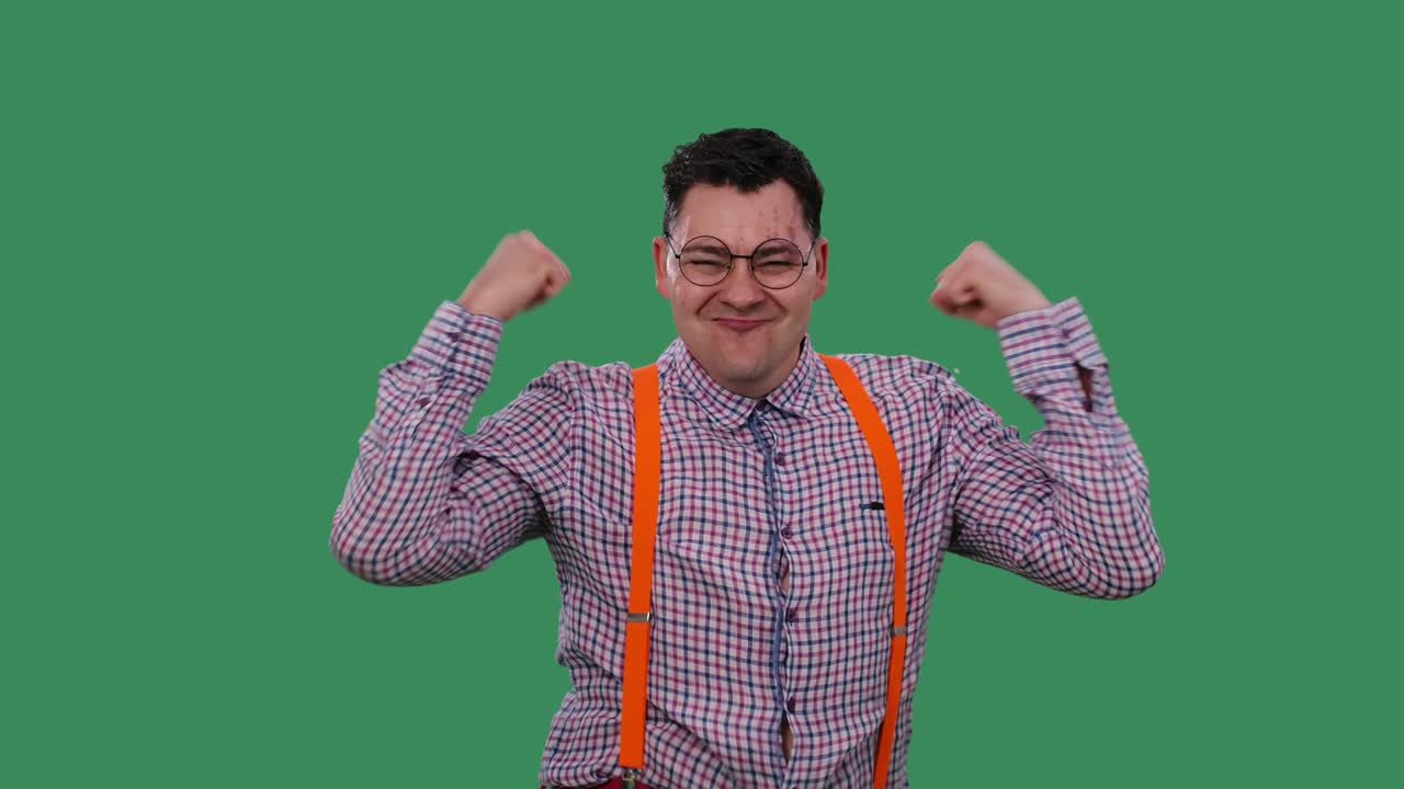 만족한 남자가 손을 들고 춤을 추고, 주먹을 고 손가락을 자기에게 가리키며 승리를 축하합니다. 녹색 스크린에 있는 스튜디오에서 안경을 쓴 남자의 초상화.