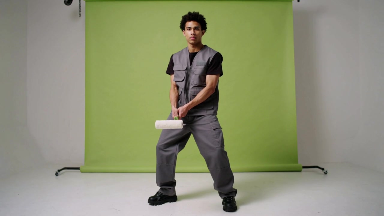 Modello uomo in movimento con gilet grigio, che tiene un rullo bianco e aggiusta la posa in studio con fondale di carta verde.