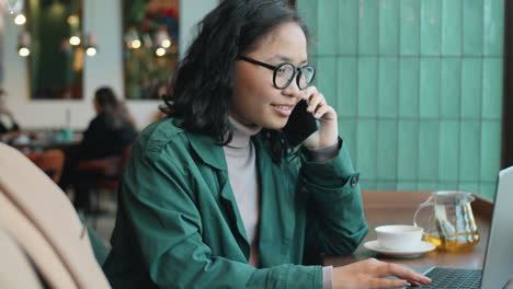 asian woman talking on phone and using laptop in cafe