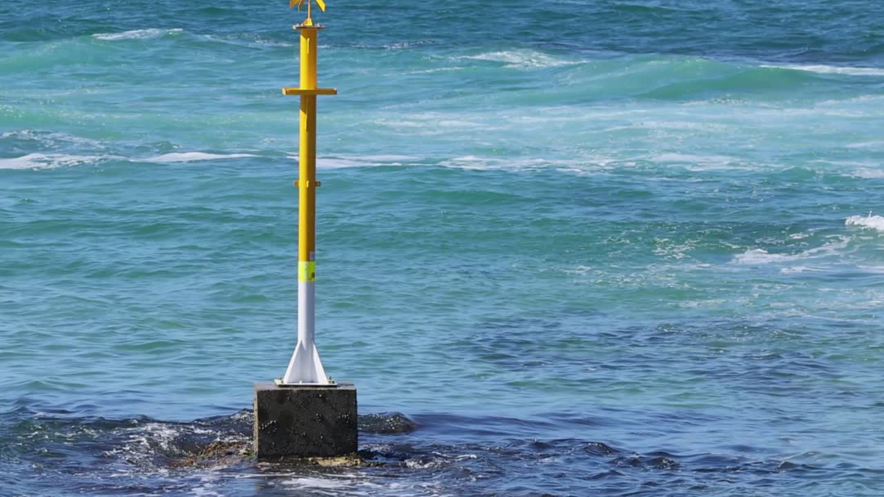 A yellow navigational marker stands firm as waves ripple and crash around it in vibrant blue waters.