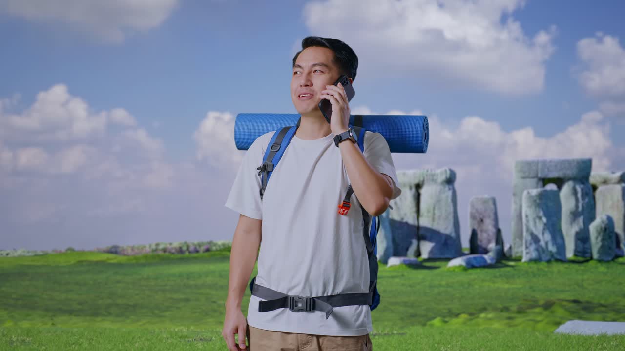 Asian Male Hiker With Mountaineering Backpack Smiling And Talking On Smartphone While Traveling In Stonehenge