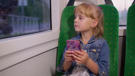 Smiling-attractive-child-girl-kid-in-city-train,-tram-or-bus-using-smartphone-chatting-with-friends