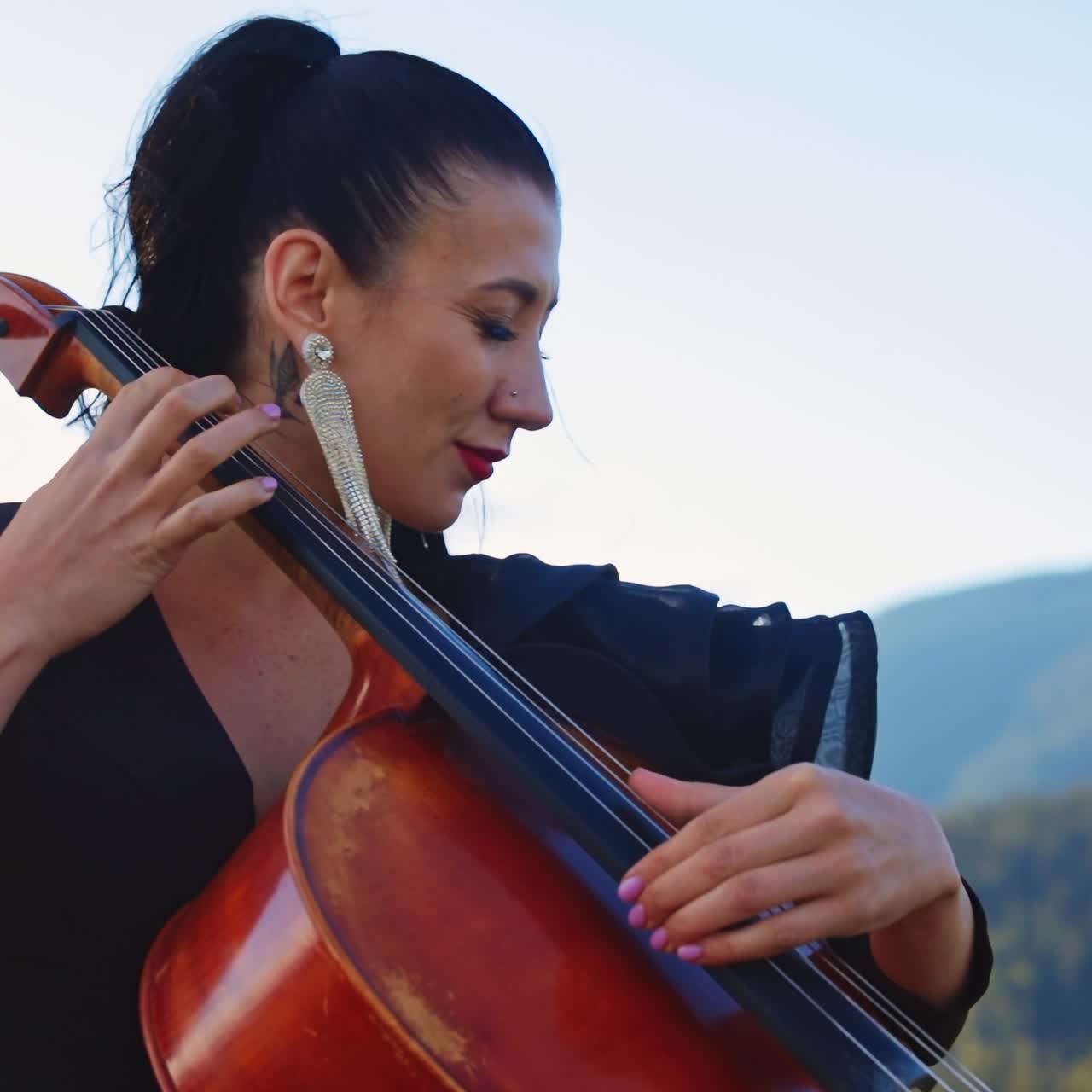 Dark-haired female musician uses no bow to play violoncello. Woman moving her head in the rhythm of music. Mountains backdrop in blur