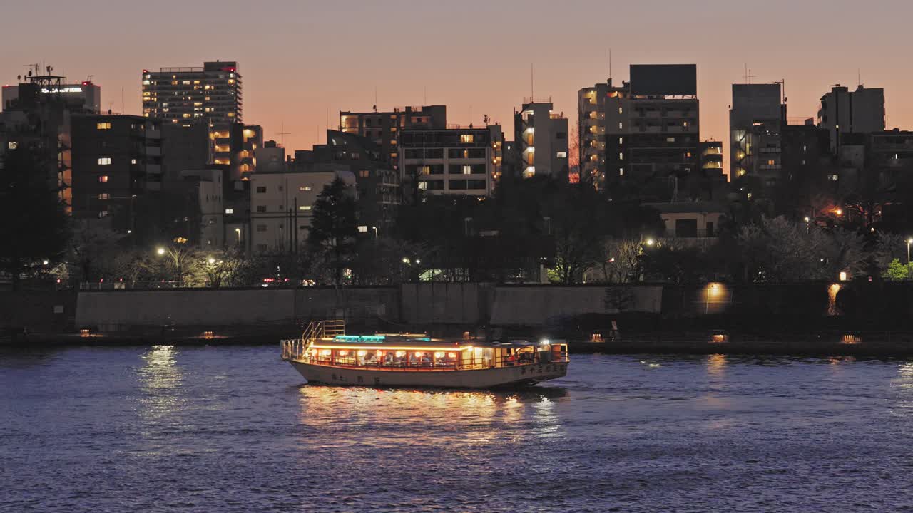 Una tradizionale barca giapponese risplende sul fiume Sumida al crepuscolo. Lo skyline della città crea uno sfondo scenografico e cinematografico in questa inquadratura fissa.