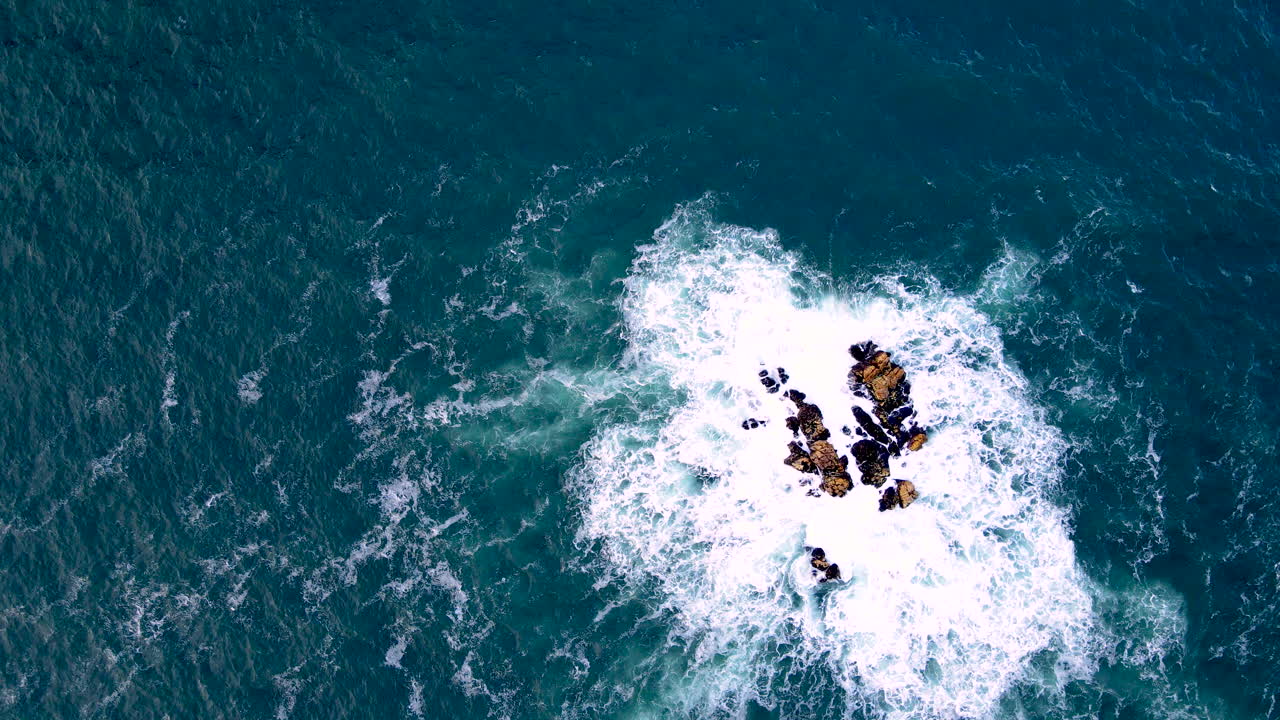 Ocean swell surging against rocky outcrop in azure blue Atlantic, top down drone