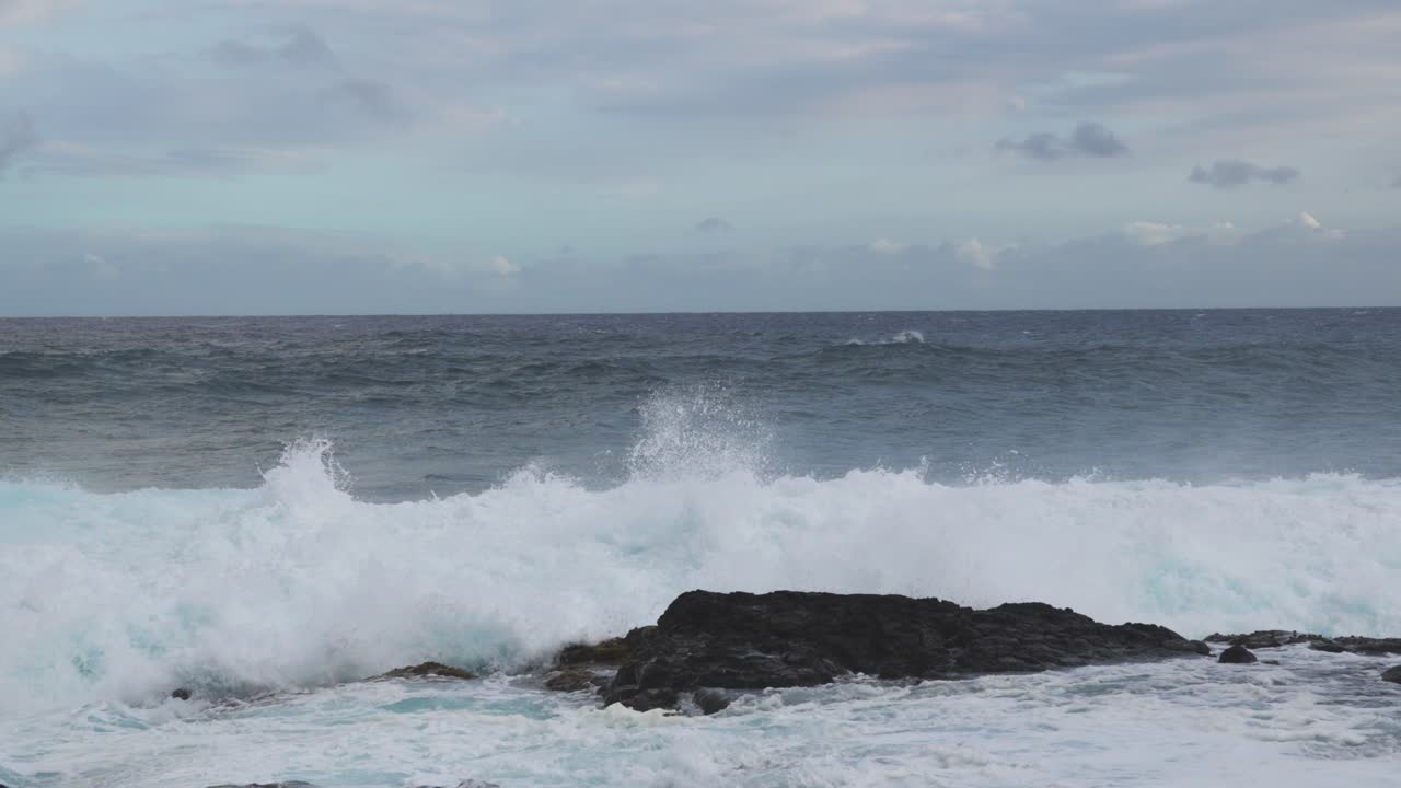 A heavy shorebreak slams into a dark rock and detonates into white spray while churning foam floods the foreground beneath pale clouds and a steady onshore breeze