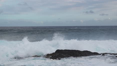 a heavy shorebreak slams into a dark rock and detonates into white spray while churning foam floods the foreground beneath pale clouds and a steady onshore breeze