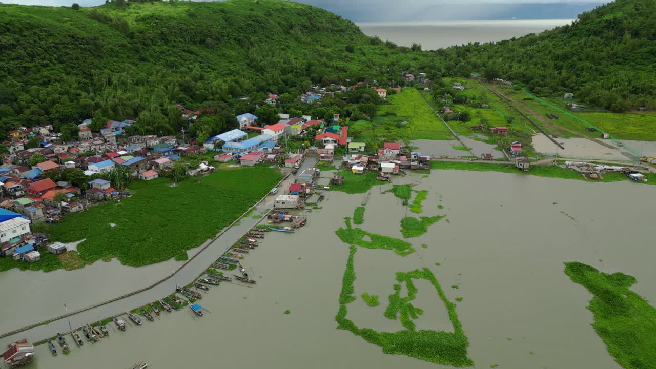 High-altitude drone shot reveals a riverside village with colorful rooftops, aquatic vegetation, and forested hills in the background