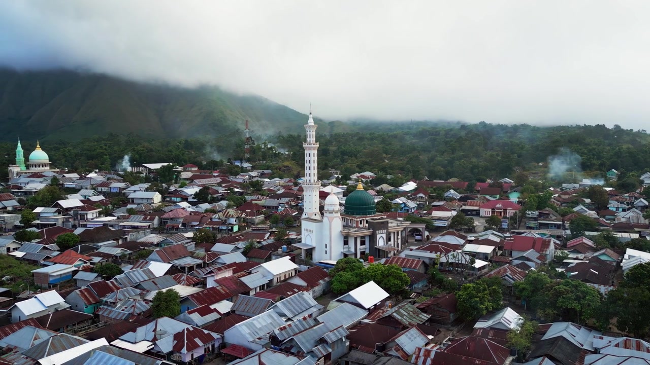 Desa pegunungan padat penduduk di bawah puncak tertutup awan dengan asap mengepul, kehidupan desa, dan masjid menonjol di pedesaan Indonesia, rekaman drone udara dengan gerakan masuk.