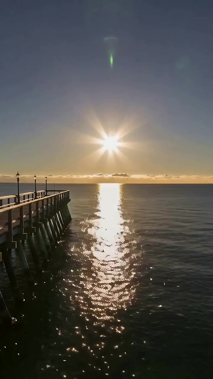 Video vertical: Sol naciente proyectando un camino brillante sobre el agua junto a los postes de luz del muelle, espacio para texto.