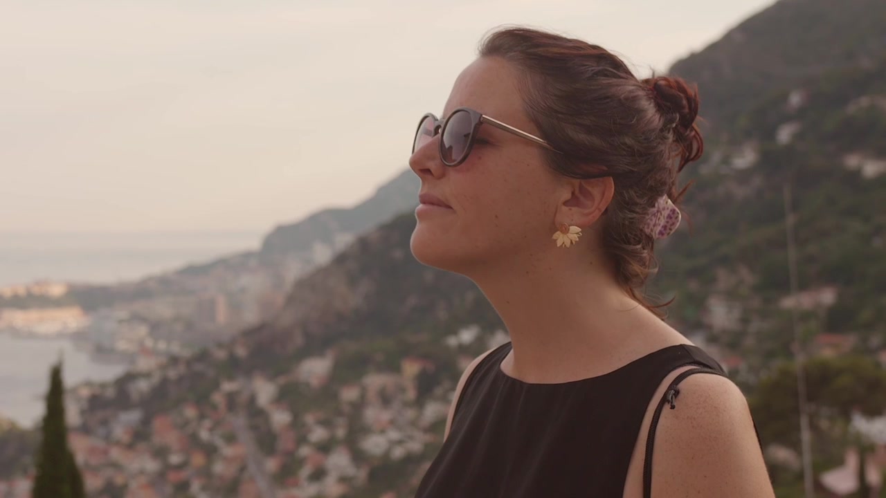 Close view of a woman wearing sunglasses while admiring the Mediterranean coastline and hillside scenery of the French Riviera.