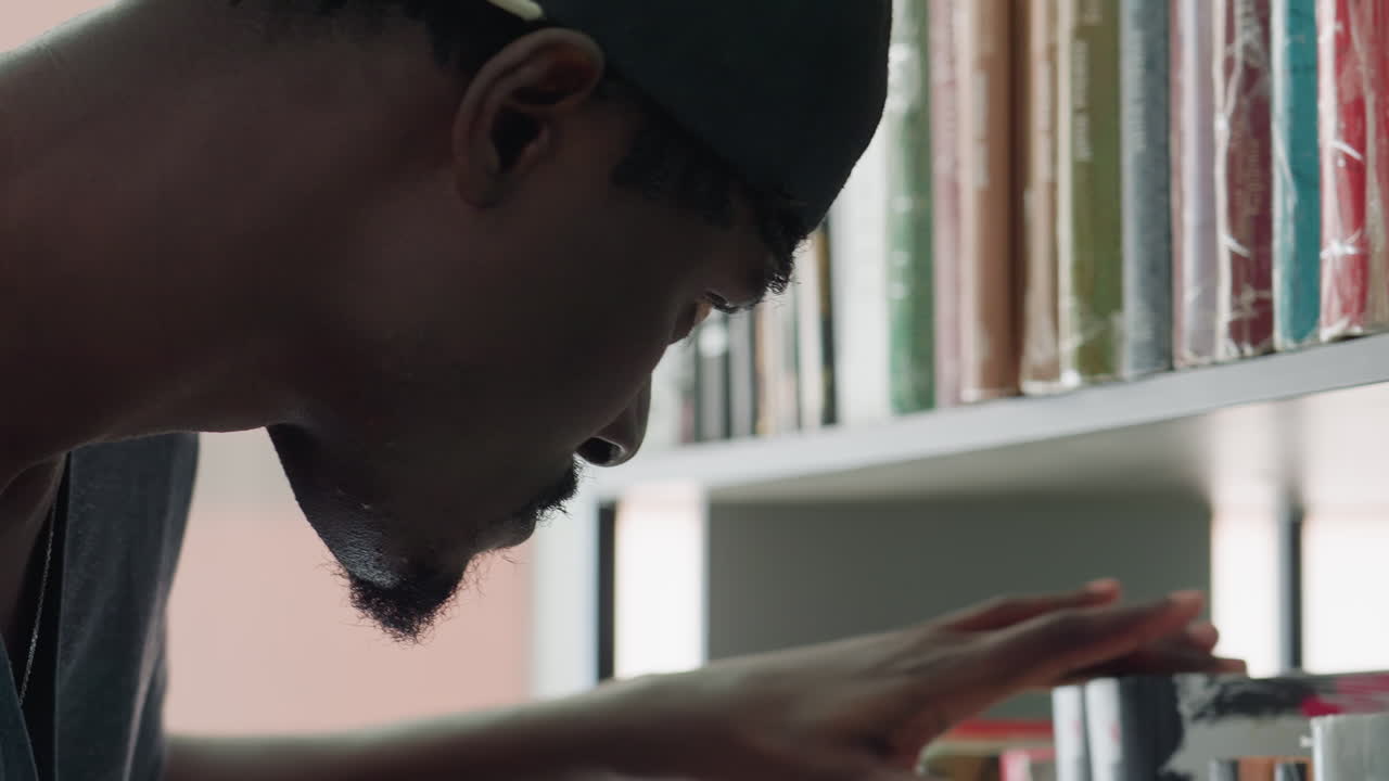 Close up of focused student wearing cap leaning toward bookshelf selecting book with concentration inside bright library setting surrounded by rows of colorful spines in academic environment