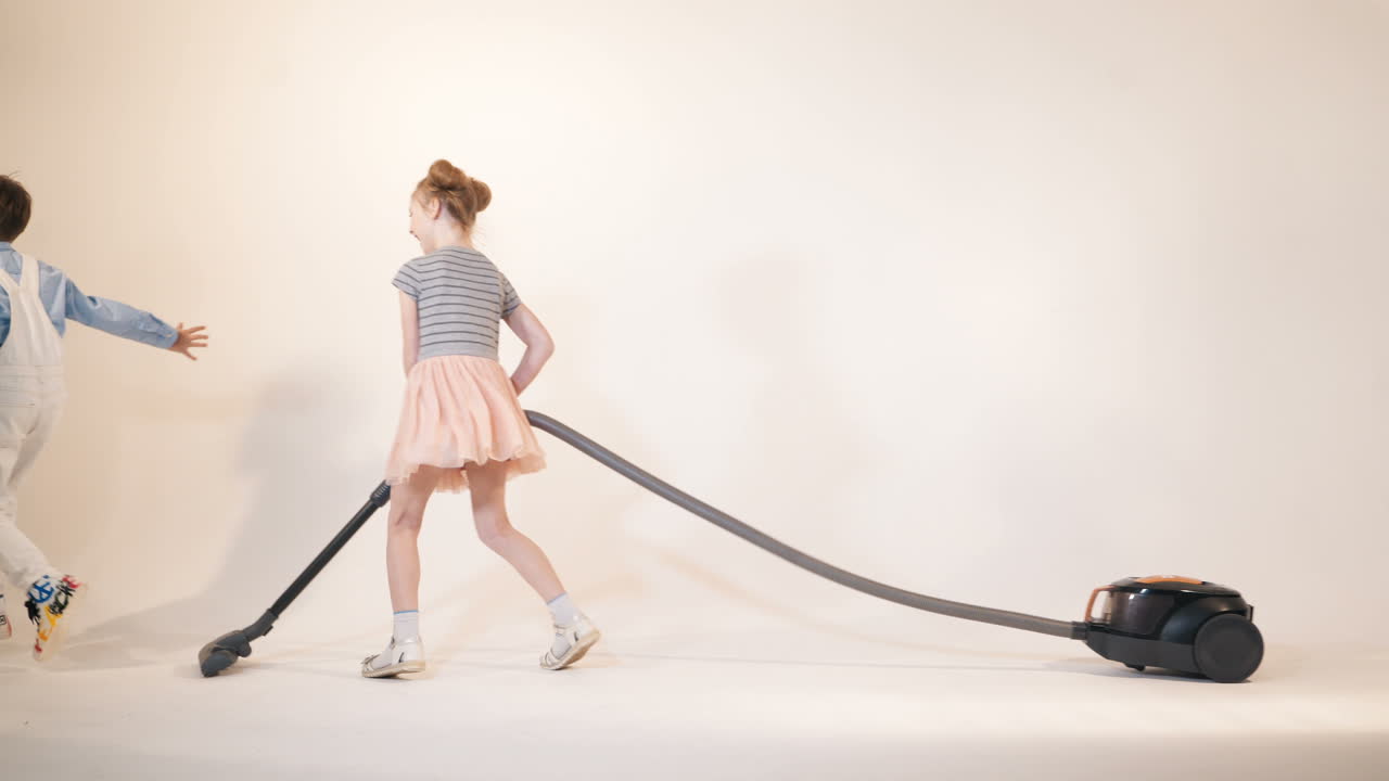 little boy is running away from the girl with a vacuum cleaner in her hands. Funny kids are playing while cleaning