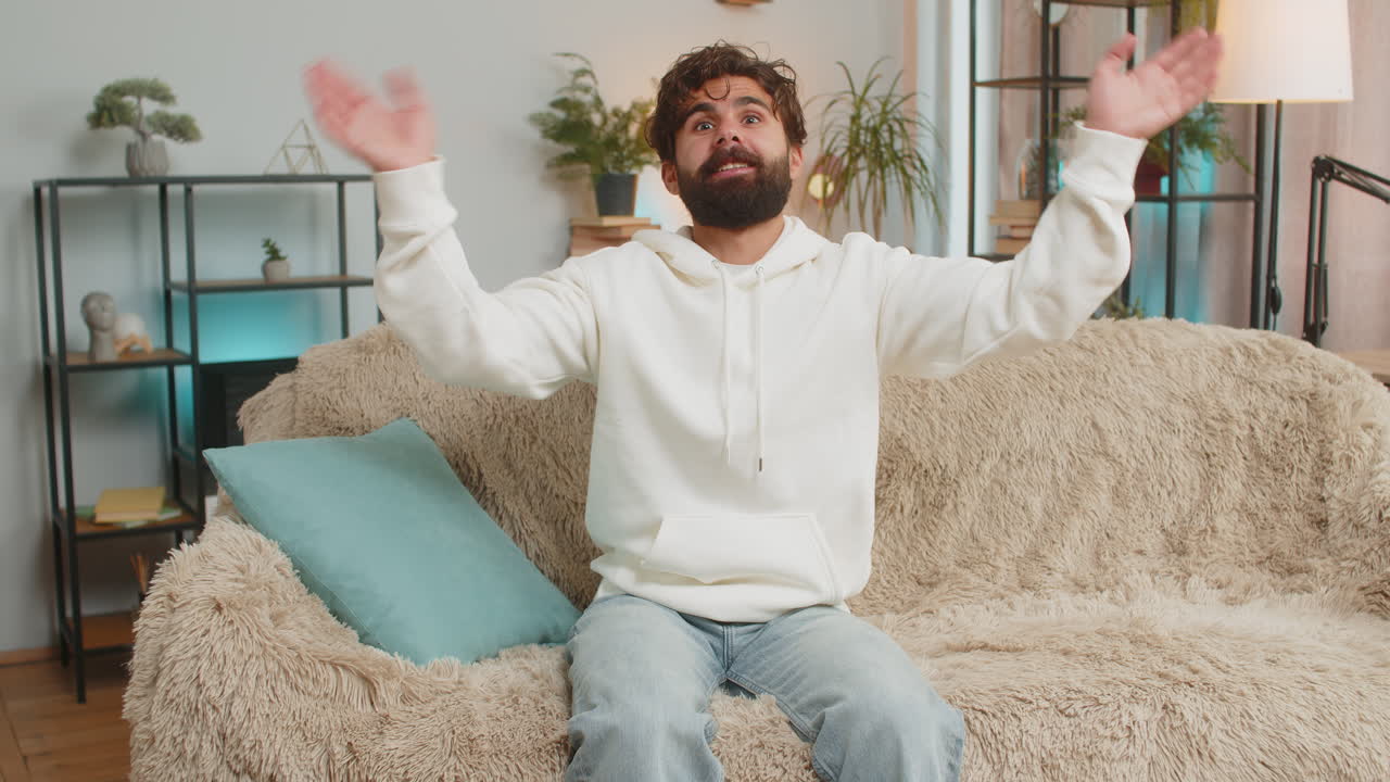 Happy young man vlogger smiling friendly at camera and waving hands while sitting on couch at home