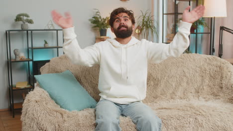 happy young man vlogger smiling friendly at camera and waving hands while sitting on couch at home