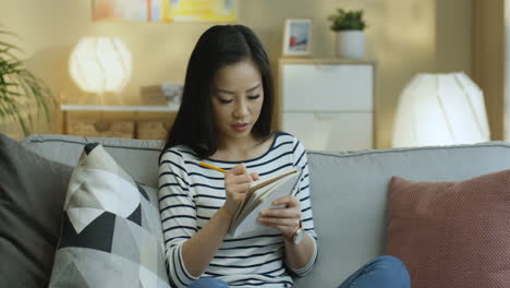 줄무늬 셔츠를 입은 젊은 여성이 공책에 메모를 하고 거실에 있는 소파에 앉아 생각하고 있다 1
