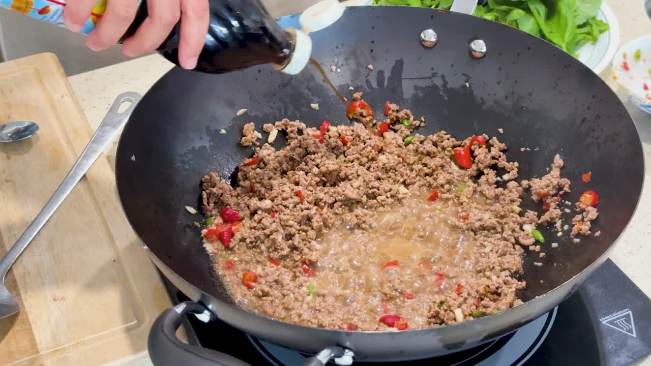 Hand pours fish sauce from a bottle into a wok with boiling ground meat and chopped vegetables, under bright kitchen lighting, top-down view