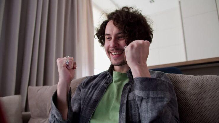 Zoom in happy brunette guy with curly hair and mustache in a checkered shirt is very happy about his success and Victory while working on a red laptop on the sofa at home