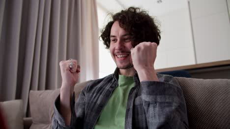 zoom in happy brunette guy with curly hair and mustache in a checkered shirt is very happy about his success and victory while working on a red laptop on the sofa at home