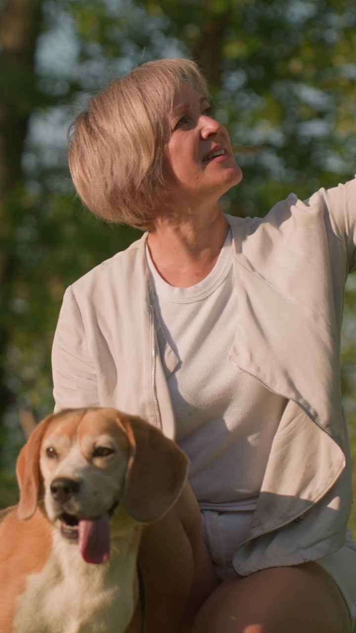 따뜻한 빛 아래 잔디에서 개와 함께 셀피를 찍는 여자, 개는 다른 것에 집중하고 주인이 나무와 초록색으로 둘러싸여 행복하게 미소 짓고 있습니다.