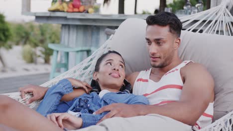 Happy-diverse-couple-relaxing-in-hammock-together-and-talking-at-beach-bar,-in-slow-motion