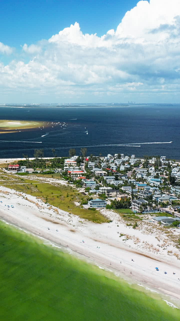 A seaside neighborhood borders a white sandy shoreline with turquoise shallows, while boats cross the dark blue horizon under a sky filled with drifting clouds