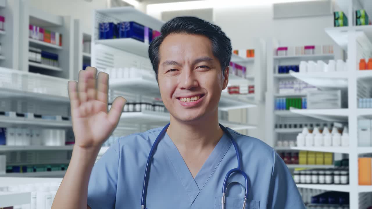 스테토스코프를 들고 있는 아시아 남성 의사가 약국에서 카메라에 손을 흔들고 미소 짓고 있습니다.