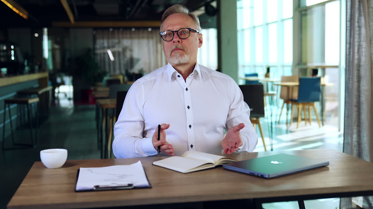 Focused senior man in white shirt sits at desk tuning the pages of his notebook. Businessman looks dissatisfied checking the information.