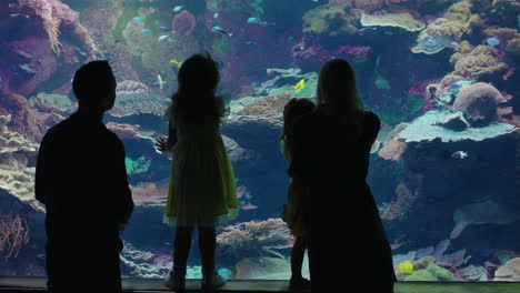 family-in-aquarium-looking-at-fish-swimming-in-corel-reef-habitat-mother-and-father-teaching-children-learning-about-marine-animals-in-oceanarium