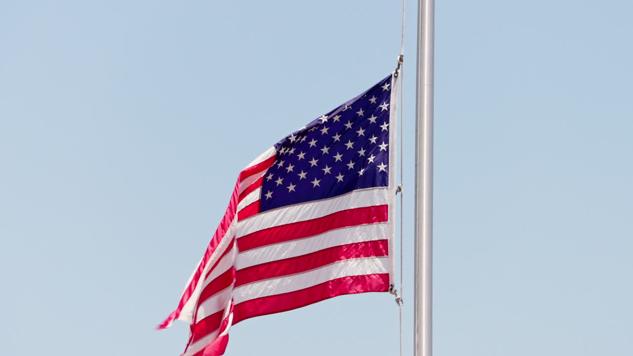 An American flag flies at half-mast in the wind, symbolizing respect and remembrance, with a clear blue sky in the background.