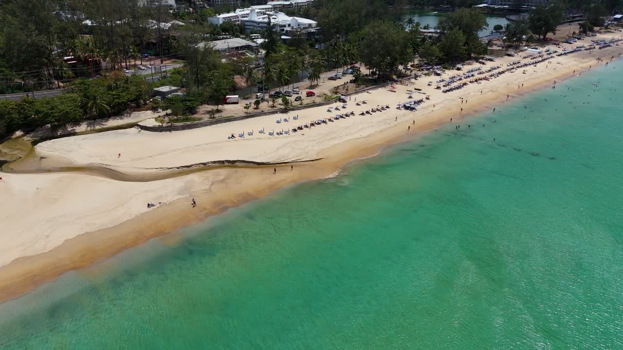 Aerial footage captures a serene beach in Phuket, Thailand, showcasing turquoise waters, sandy shores, and sunbathers under clear skies